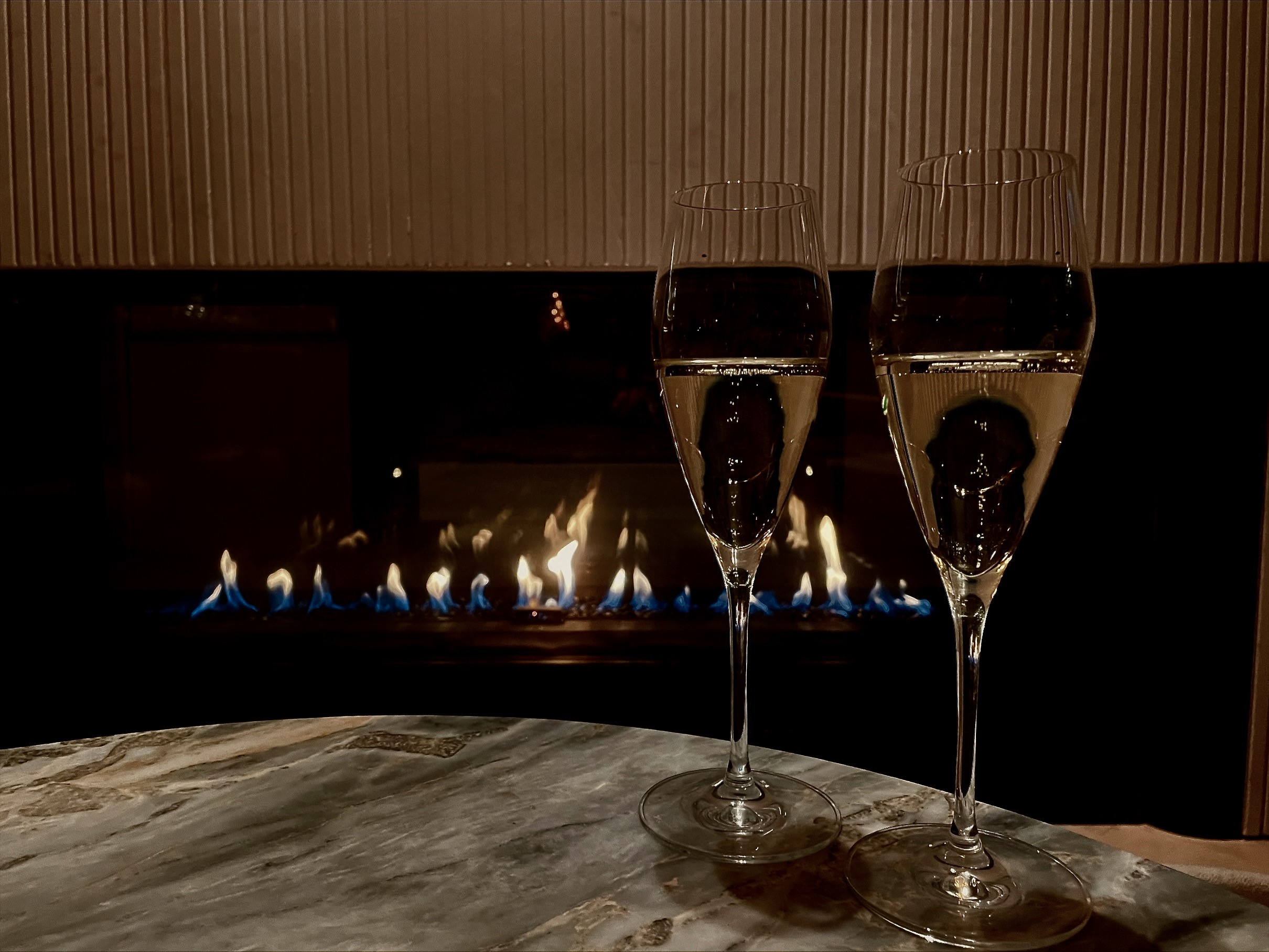Two glasses of champagne sitting on a table by the warm fire.