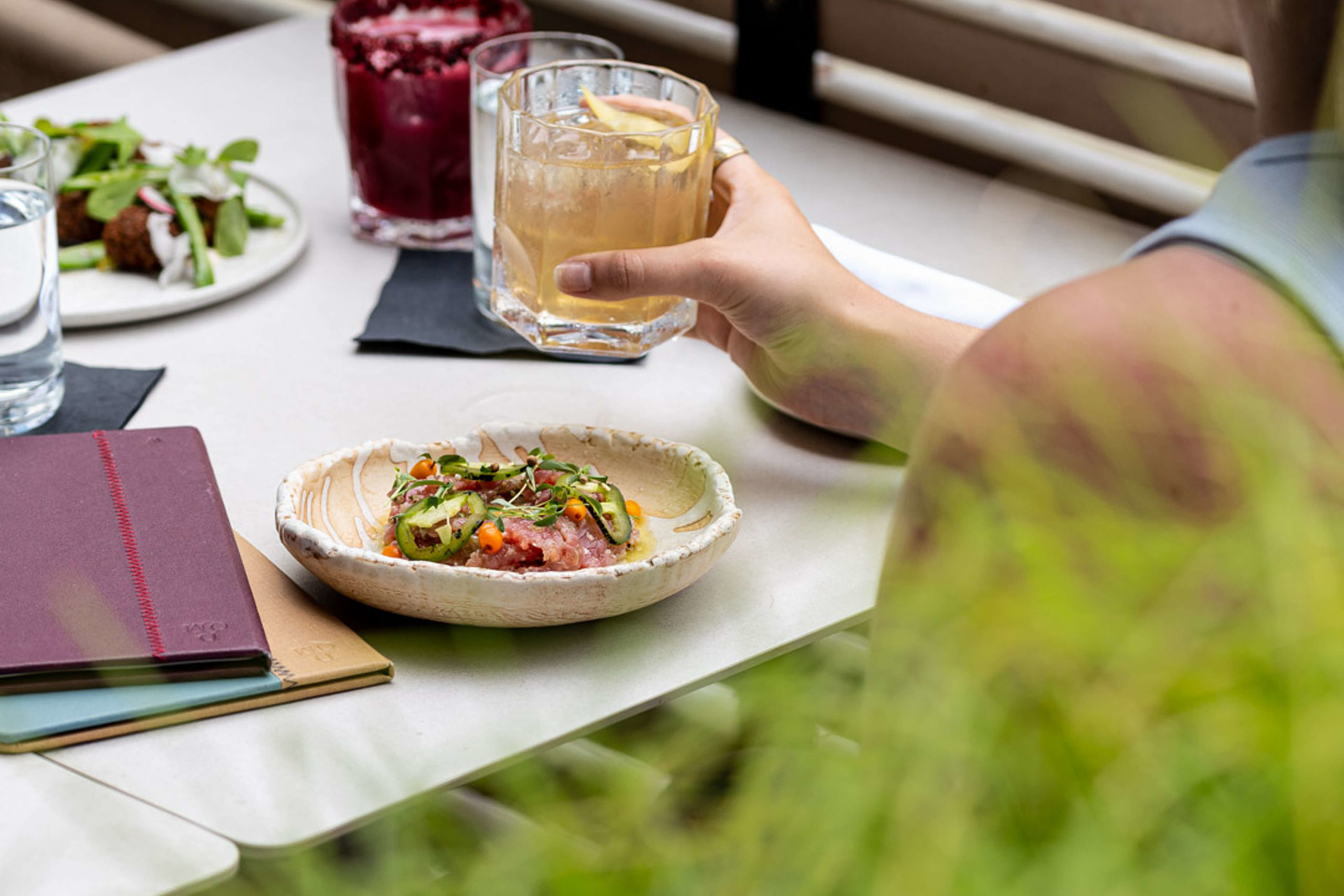 Woman holds drink sitting at patio table with TUNA SASHIMI 19 sea buckthorn, charred jalapeño Drift Hailifax